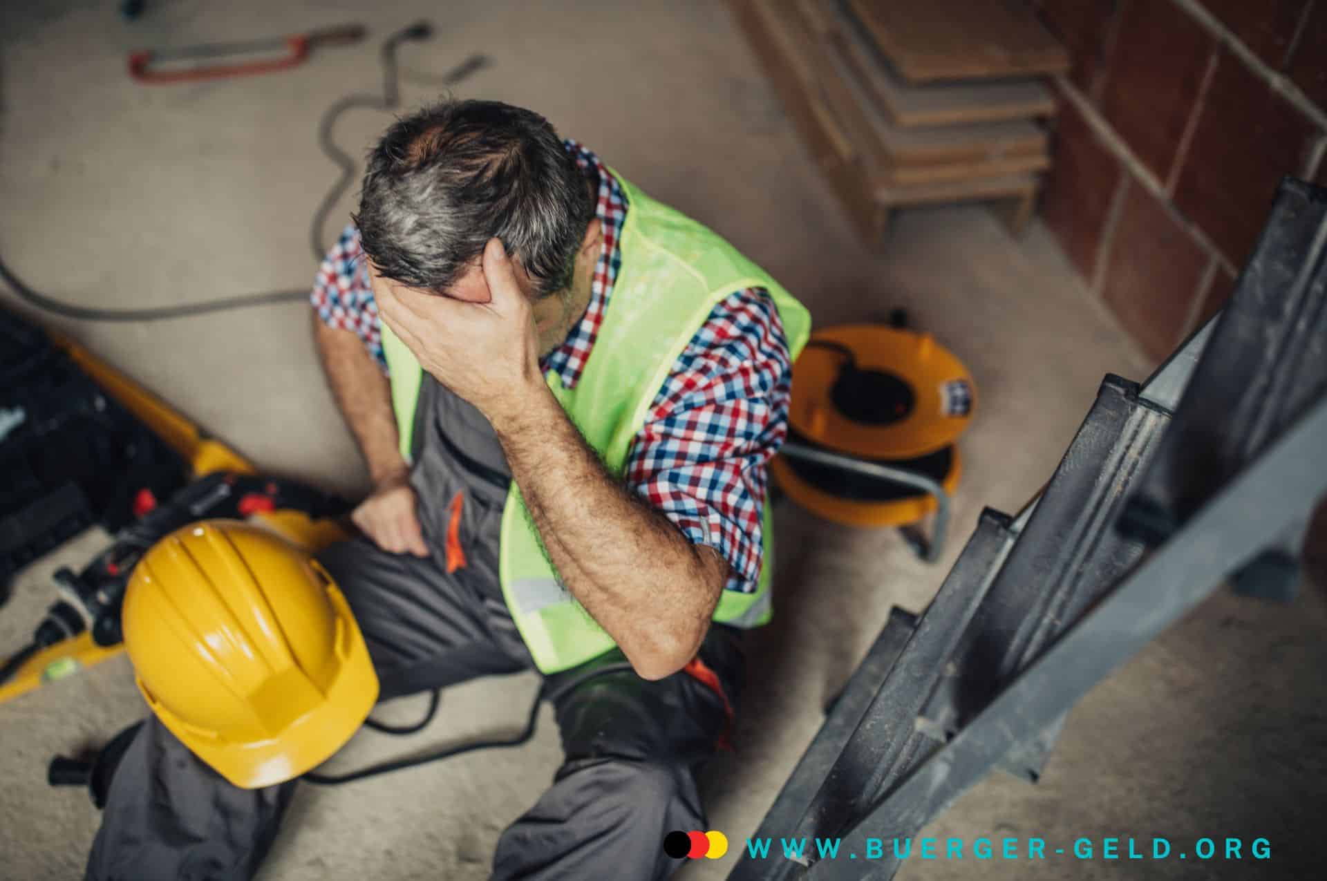 Arbeiter mit Helm sitzt auf dem Boden und hält sich die Stirn