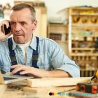 Älterer Mann sitzt am Computer in Werkstatt und telefoniert.